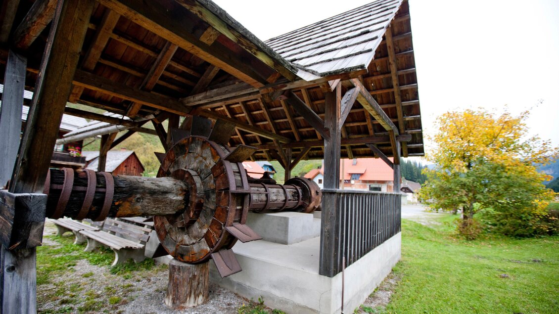 Eine historische Mühle mit einem großen Holzrad steht unter einem Dach aus Holzschindeln. Umgeben von grünem Gras und ein paar Bäumen im Hintergrund. | © Holzwelt Murau
