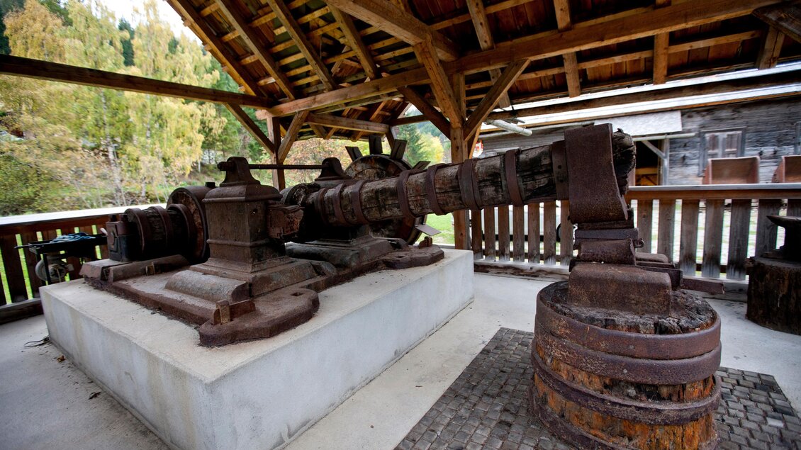 Eine alte, rustikale Maschine steht unter einem überdachten Bereich. Die Maschine hat einen robusten Metallrahmen und ist auf einem weißen Betonfundament installiert. | © Holzwelt Murau