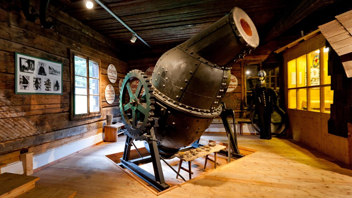 Ein historisches Gerät in einem Museum, das an eine große, schwarze Maschine erinnert. Der Raum hat eine rustikale Holzdecke und einige Bilder an den Wänden. | © Holzwelt Murau