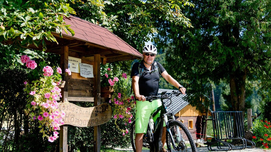 Ein Mann steht an einem Holzschild mit Blumen im Hintergrund und hält ein Fahrrad. Die Szenerie zeigt eine grüne, bewaldete Landschaft. | © TV Region Graz - Mias Photoart