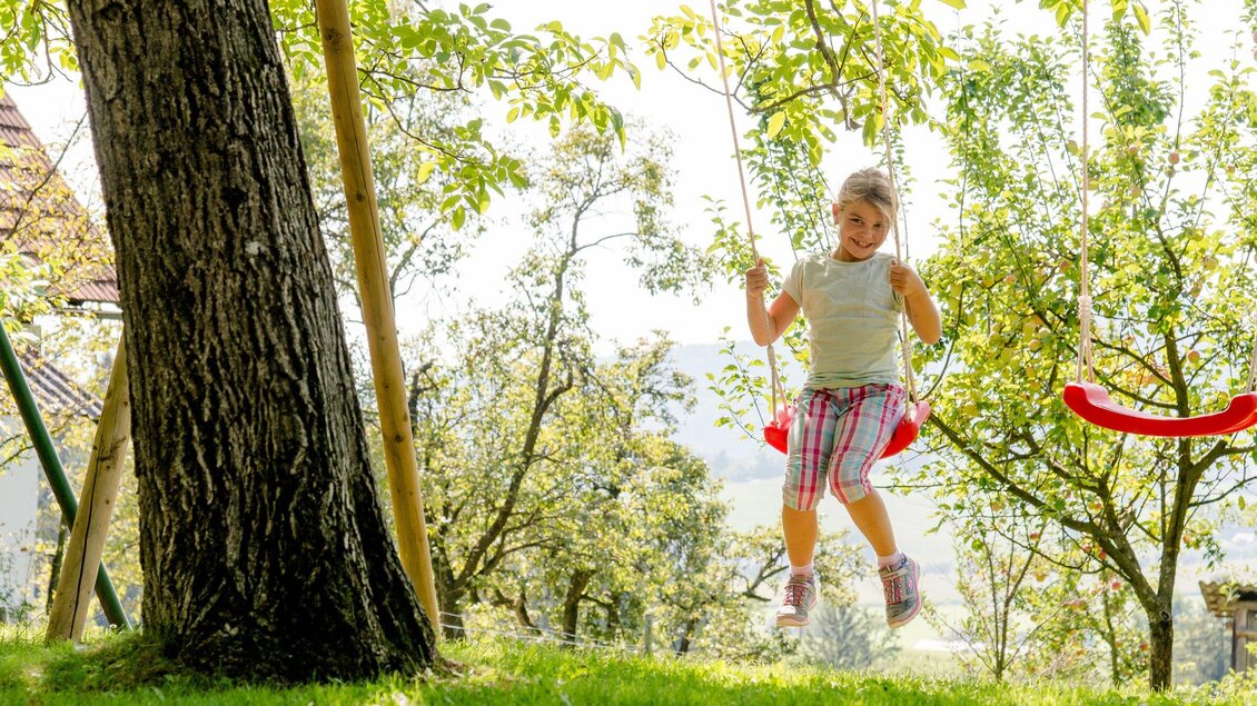 Ein Mädchen schwingt fröhlich auf einer Schaukel im Garten. Umgeben von Bäumen genießt sie den Sonnenschein. | © TV Region Graz - Mias Photoart