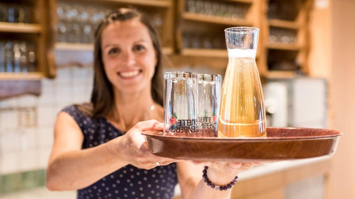Eine lächelnde Frau hält ein Tablett mit einem Krug und Gläsern. Im Hintergrund sind Regale mit Getränken zu sehen. | © Serschenhof