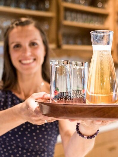 A smiling woman is holding a tray with a jug and glasses. In the background, shelves with drinks are visible. | © Serschenhof