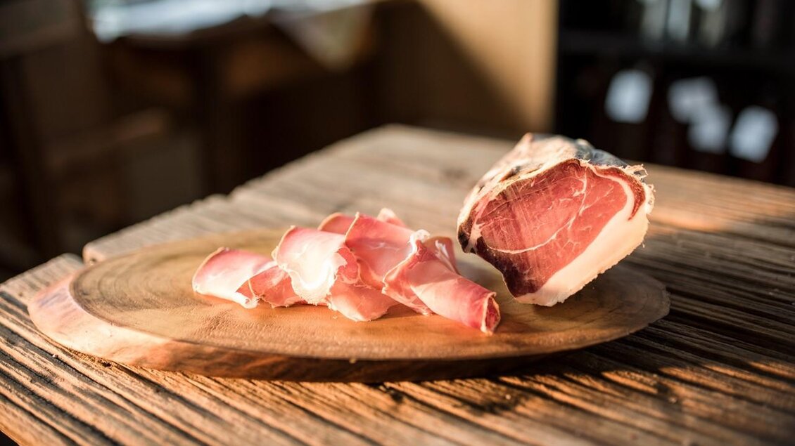 Rohschinken auf einem Holzbrett, mit einigen dünnen Scheiben daneben. Die Oberfläche ist rustikal und warm beleuchtet. | © Serschenhof