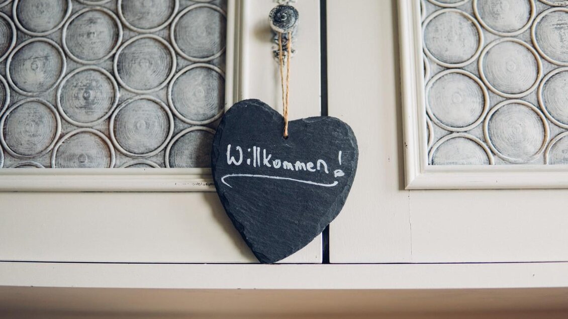 Ein herzförmiges Schild aus Schiefer hängt an einem Schrank. Darauf steht „Willkommen!“ in weißer Schrift. | © Serschenhof