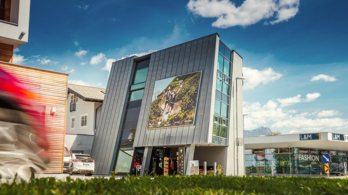 Ein modernes Gebäude mit großen Fenstern und einer Werbetafel an der Fassade. Im Vordergrund sieht man eine grüne Wiese und blauen Himmel mit Wolken. | © Mountain Shop Schladming