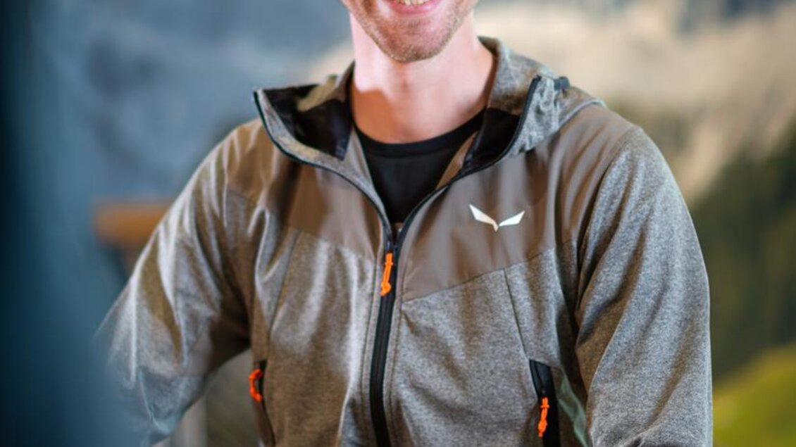 Ein junger Mann mit einem freundlichen Lächeln sitzt in sportlicher Kleidung. Im Hintergrund sind Berge und eine natürliche Umgebung zu sehen. | © Mountain Shop Schladming