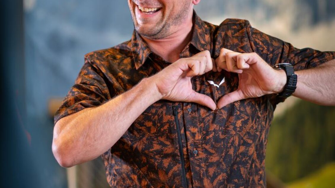 Ein lächelnder Mann macht ein Herz mit seinen Händen. Im Hintergrund sind Berge zu sehen. | © Mountain Shop Schladming