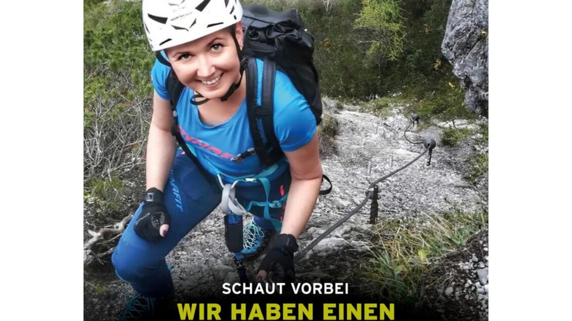 Eine Frau in einer Kletterausrüstung, die an einer Felswand klettert. Der Text bewirbt einen Verleih von Klettersteigsets in einem Bergshop. | © Salewa Mountain Shop Schladming