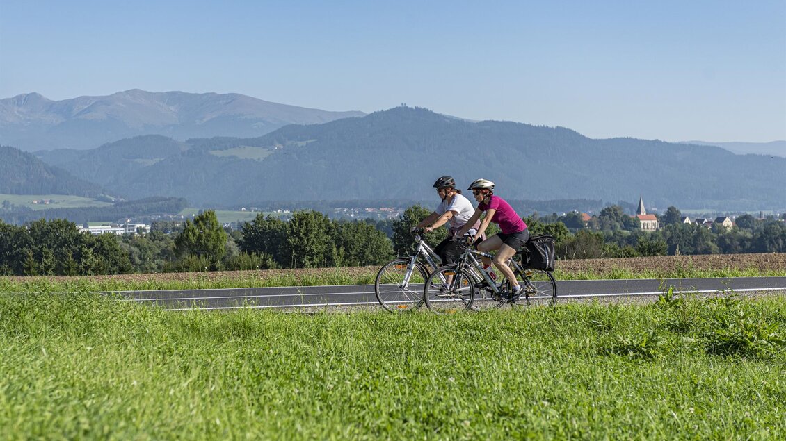 Zwei Radfahrer fahren auf einer Straße inmitten einer grünen Landschaft. Im Hintergrund sind Hügel und ein blauer Himmel zu sehen. | © Anita Fössl