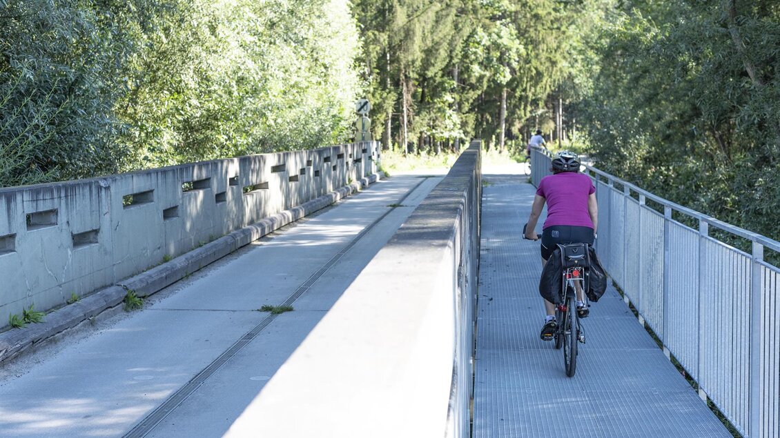Eine Radfahrerin fährt über eine Brücke in einer grünen Umgebung. Die Straße ist breit und von Bäumen umgeben. | © Anita Fössl