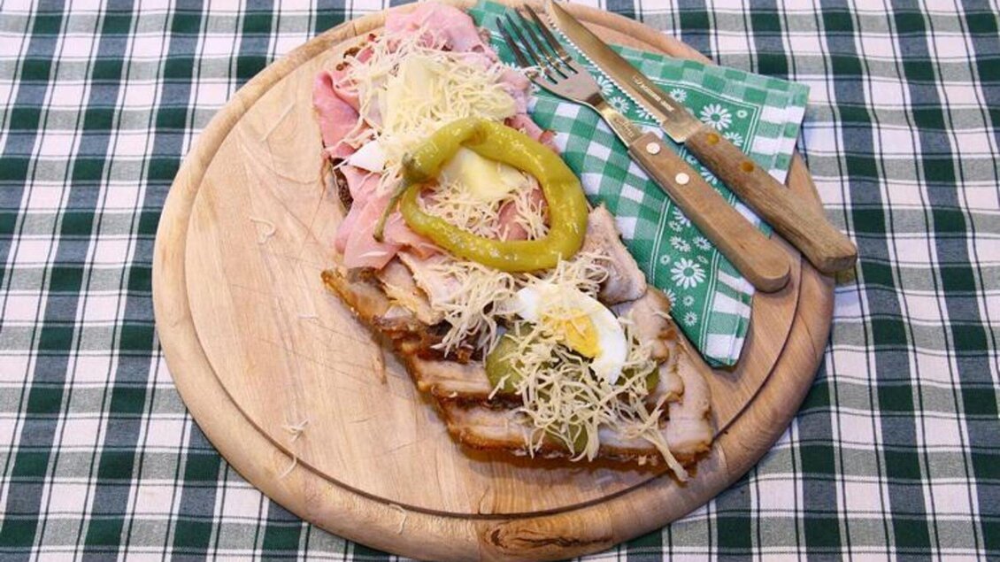 Ein Holzbrett mit einem großen belegten Brot, garniert mit Schinken, Käse, Oliven und einer Gurkenscheibe. Daneben liegen eine Gabel und ein Messer auf einem karierten Tischtuch. | © Mur Heuriger