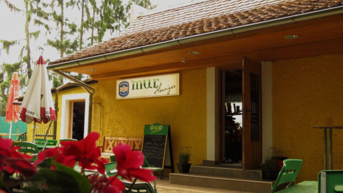 Ein einladendes Restaurant mit gelber Fassade und einem Eingang. Im Vordergrund sind Blüten und grüne Stühle sichtbar. | © Mur Heuriger