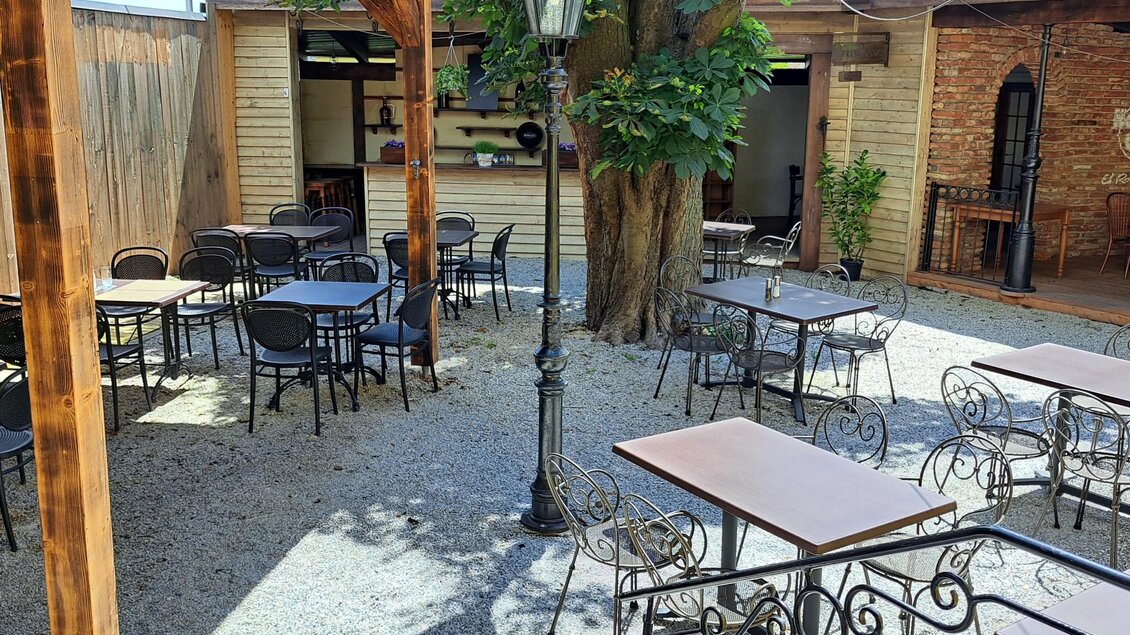 Ein gemütlicher Außenbereich mit schwarzem Bistro-Mobiliar und einem großen Baum in der Mitte. Eine Laterne steht zwischen den Tischen und schafft eine einladende Atmosphäre. | © Johannes Ulrich