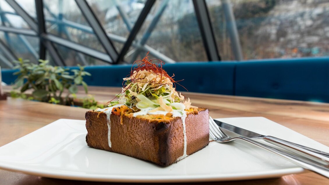 Ein kunstvoll angerichtetes Gericht auf einem weißen Teller. Es besteht aus einem Stück Brot, garniert mit frischem Gemüse und einer cremigen Sauce. | © Murinsel Café