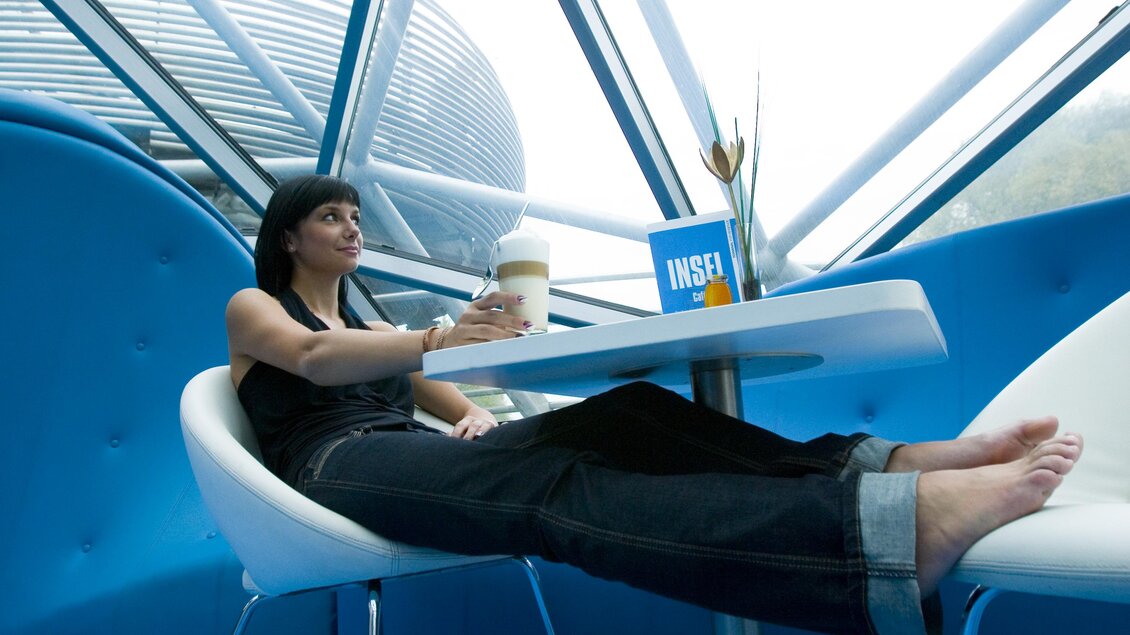 Frau sitzt barfuß mit Latte Macchiato an einem Tisch im modernen, blau gestalteten Innenbereich der Murinsel. | © Graz Tourismus - Tom Lamm