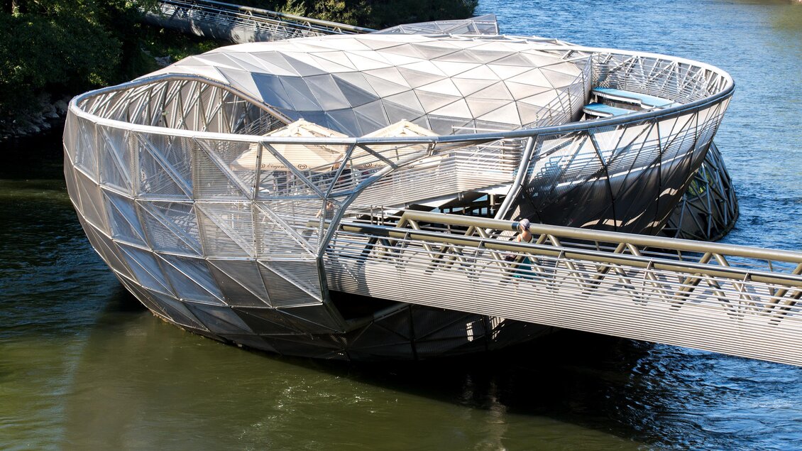 Ein modernes, gläsernes Gebäude am Fluss mit einer einzigartigen, schalenartigen Form. Eine Brücke führt zu dem Bauwerk, umgeben von grünen Bäumen und Wasser. | © Graz Tourismus - Harry Schiffer