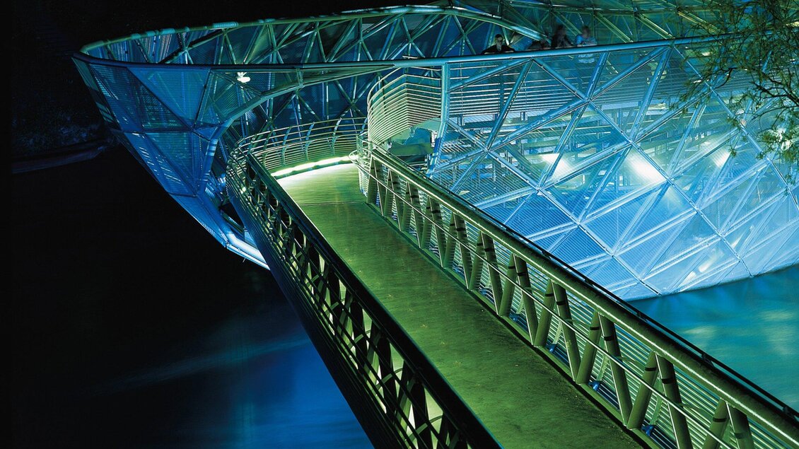 Eine moderne Brücke aus Glas und Stahl über das Wasser. Sie ist in sanftem Licht beleuchtet und hat eine geschwungene Form. | © Graz Tourismus - Harry Schiffer