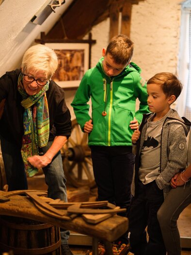 Eine Gruppe von Kindern schaut einer älteren Dame zu, während sie etwas erklärt. Sie befinden sich in einem rustikalen Raum mit historischen Werkzeugen und Möbeln. | © Kump.Photography