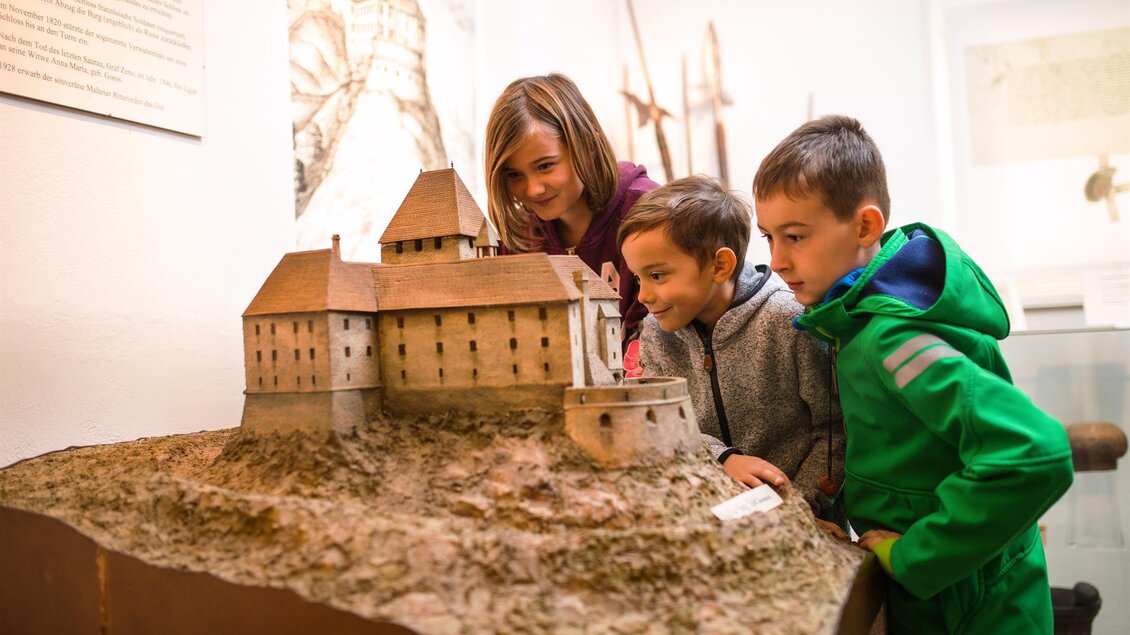 Drei Kinder betrachten eine detaillierte Modellburg in einem Museum. Die Kinder sind fasziniert und lächeln, während sie das Modell erkunden. | © Kump.Photography