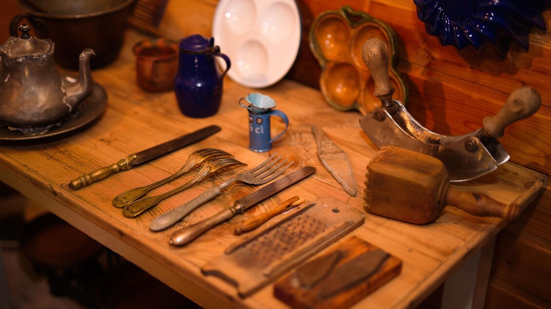 Ein Holztisch mit verschiedenen alten Küchenutensilien, darunter Besteck und Töpfe. Im Hintergrund sind weitere Geschirrteile und eine blaue Wanddekoration zu sehen. | © Kump.Photography