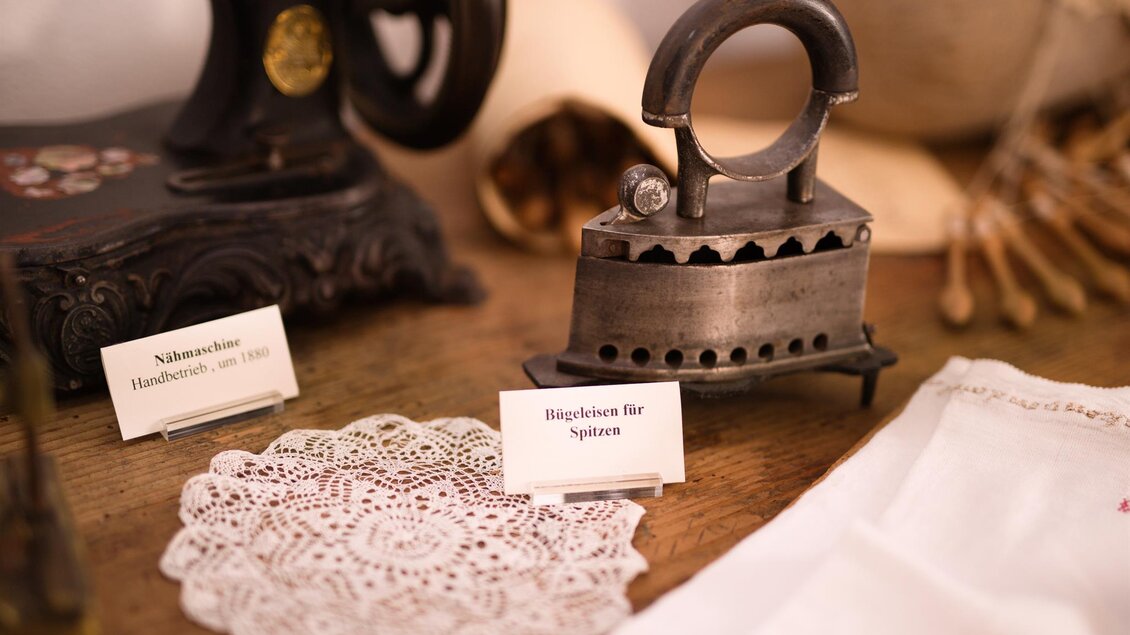 Eine alte Nähmaschine steht neben einem nostalgischen Bügeleisen und einer filigranen Tischdecke. Auf dem Tisch befinden sich auch Beschriftungen mit Informationen zu den Objekten. | © Kump.Photography