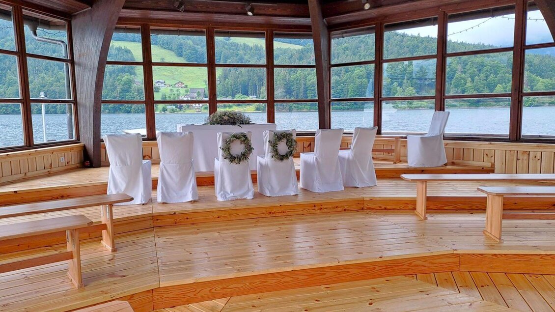 Ein gemütlicher Veranstaltungsraum mit großen Fenstern und Blick auf einen See. Der Raum ist mit weißen Stühlen und einem dekorierten Tisch ausgestattet. | © Gemeinde Grundlsee, Theresa Grill
