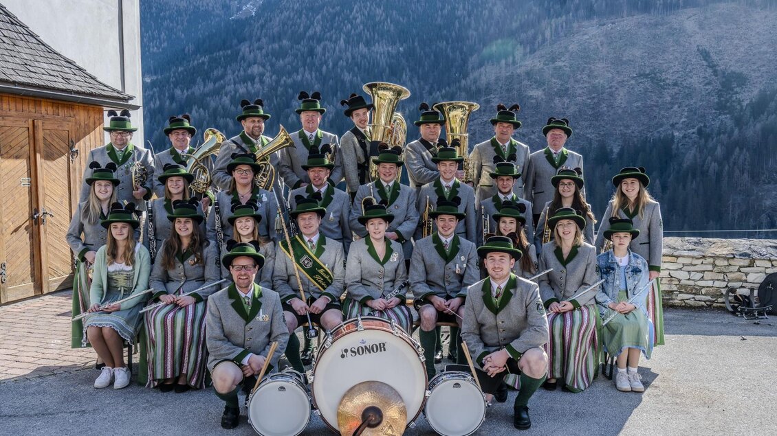 Eine Musikkapelle steht in Trachtenkleidung vor einer Berglandschaft. Die Mitglieder lächeln und posieren für das Foto. | © Heinz & Melitta Holzmüller