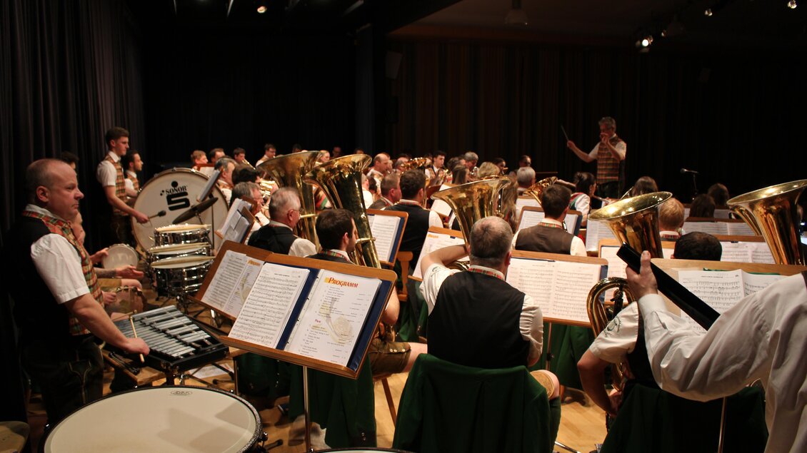 Eine große Blaskapelle spielt auf der Bühne. Der Dirigent leitet die Musiker und es sind mehrere Instrumente zu sehen.