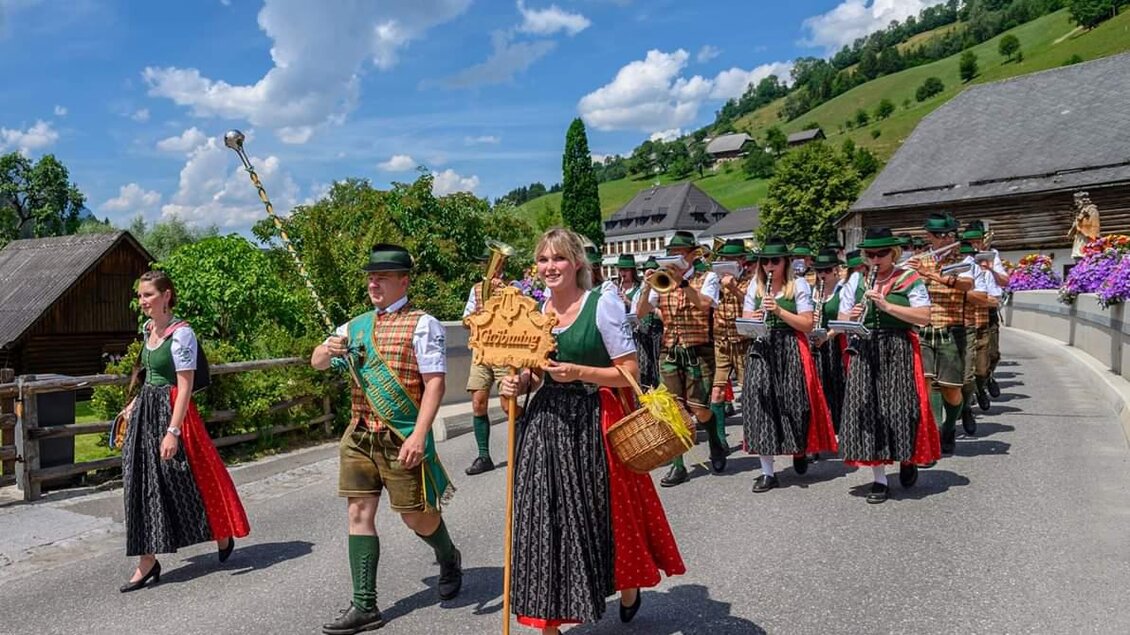 Eine Gruppe von Menschen in traditioneller Tracht marschiert eine Straße entlang. Im Hintergrund sind grüne Hügel und Blumenkästen zu sehen.