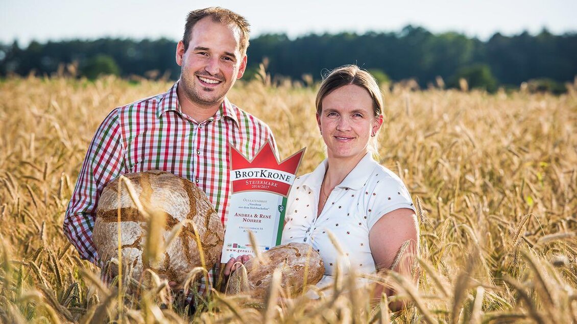 Ein Paar steht in einem Weizenfeld und hält frisch gebackenes Brot sowie eine Auszeichnung in den Händen. Die Sonne scheint und die Atmosphäre ist freundlich und einladend. | © Nöhrer
