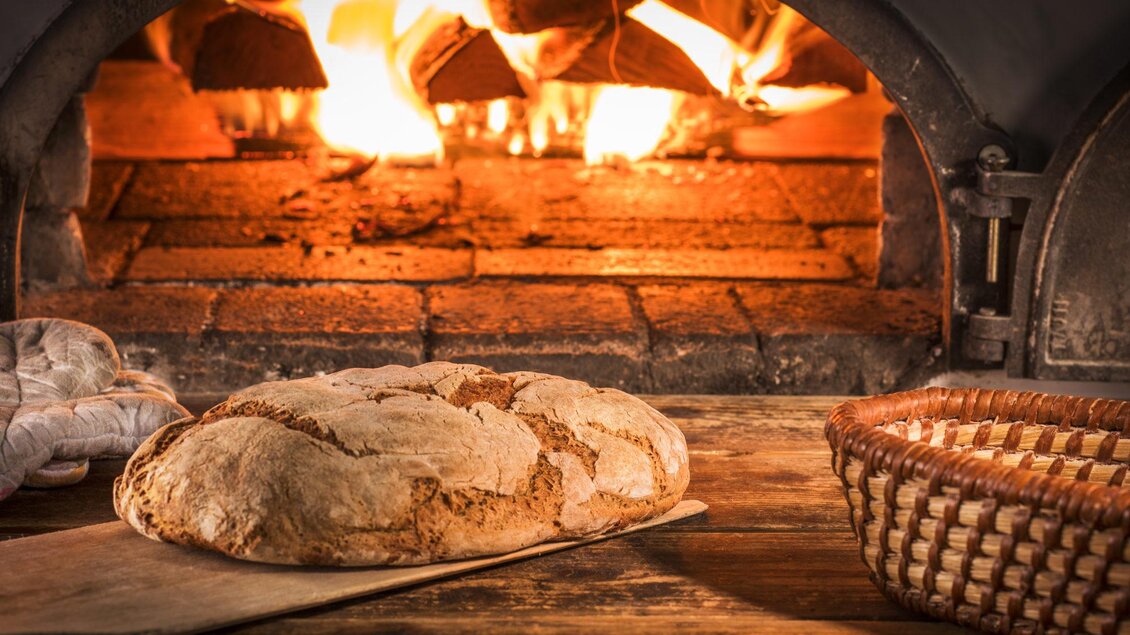 Ein frisch gebackenes Brot liegt auf einem Holzbrett vor einem warmen Ofen. Im Hintergrund brennen die Flammen und erzeugen eine gemütliche Atmosphäre. | © Nöhrer