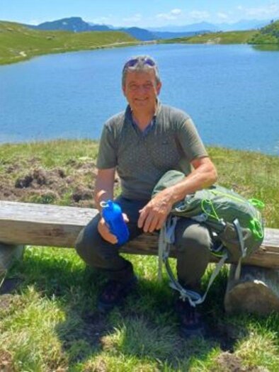 A man is sitting on a bench by the shore of a clear lake. In the background, green hills and blue sky can be seen. | ©  Matthias Pointinger
