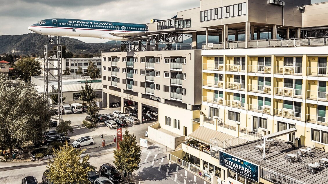 Ein modernes Hotel mit einem Flugzeugmodell auf dem Dach. Die Umgebung ist urban mit Parkplätzen und Bäumen. | © Novapark - Ulrike Rauch