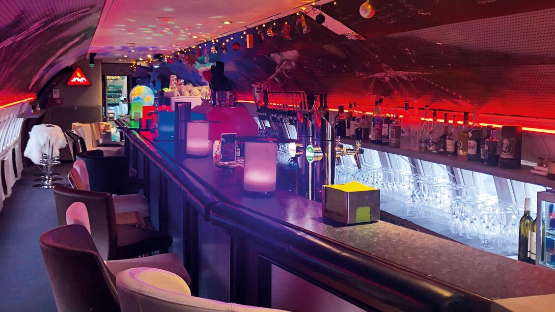 Eine moderne Bar mit stilvollem Design und bunten Lichtern. Der lange Bartresen ist mit verschiedenen Getränken und Gläsern dekoriert. | © NOVAPARK