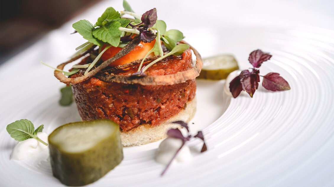 Ein kunstvoll angerichtetes Gericht mit einer gemischten Fleischmasse, garniert mit frischen Kräutern und kleinen Gurkenscheiben. Die Präsentation erfolgt auf einem eleganten weißen Teller. | © NOVAPARK - Ulrike Rauch