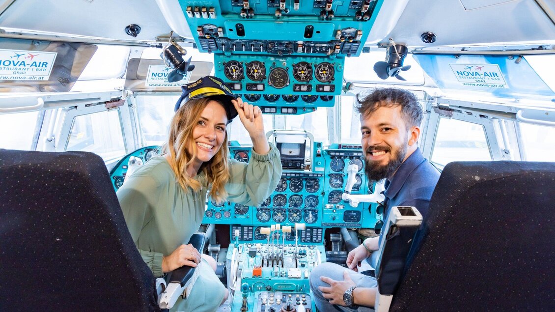 Zwei Personen sitzen im Cockpit eines Flugzeugs. Die Frau trägt eine Pilotenmütze und lächelt, während der Mann ebenfalls lächelt. | © Graz Tourismus - Werner Krug