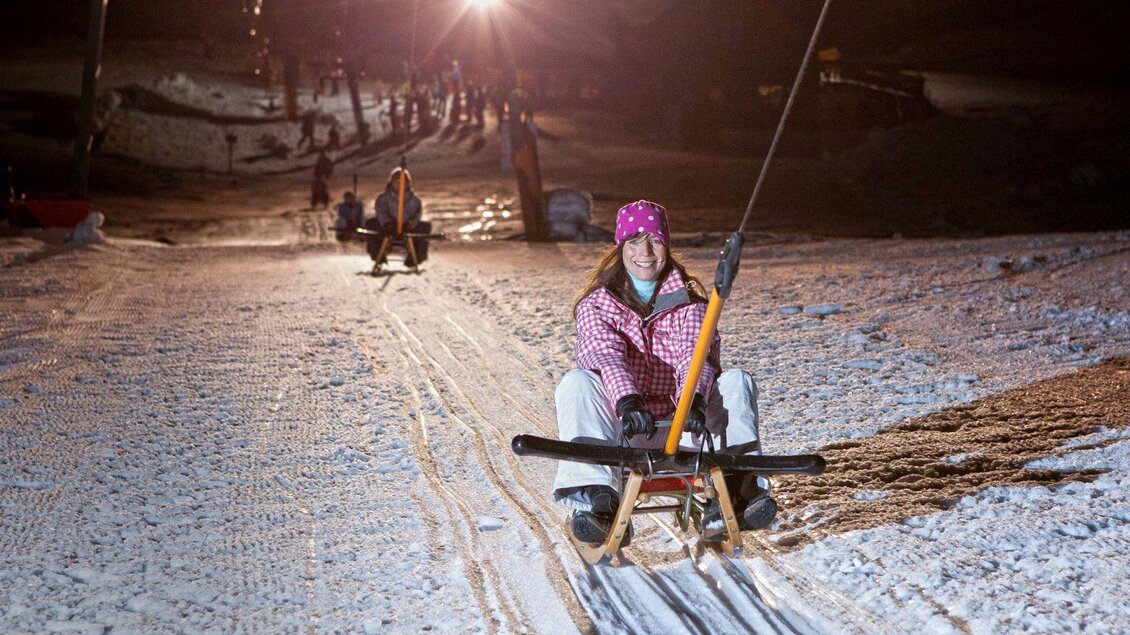 Eine Frau sitzt auf einem Schlitten und fährt bei Nacht auf einer beleuchteten Piste. Im Hintergrund sind andere Rodler und Schneelandschaft sichtbar. | © Mirja Geh