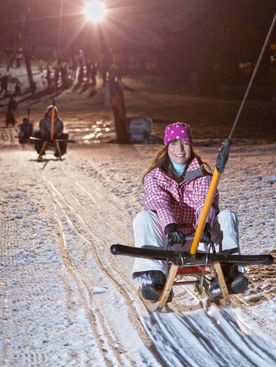 A woman is sitting on a sled and riding at night on a lit slope. In the background, other sledders and a snowy landscape are visible. | © Mirja Geh