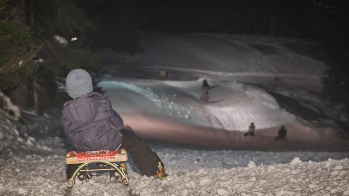 Ein Kind sitzt auf einem Schlitten und blickt auf einen verschneiten Hang. Im Hintergrund sind mehrere Personen, die den Hang hinunter rutschen.