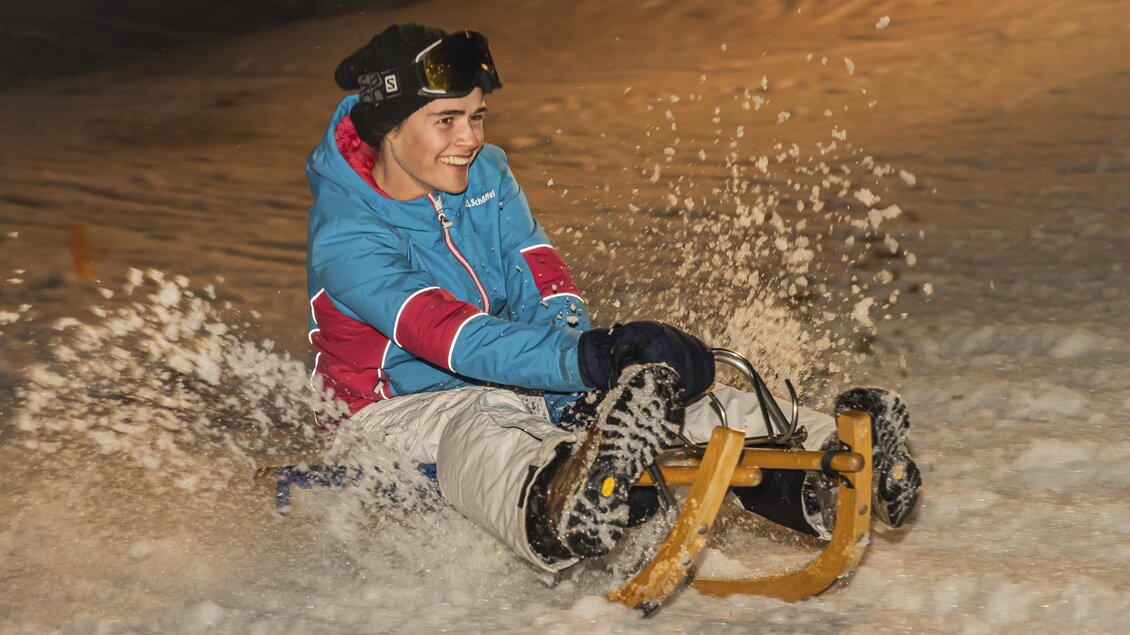 Ein Junge fährt mit einem Schlitten den verschneiten Hang hinunter. Der Abendhimmel ist leicht beleuchtet und Schnee spritzt auf.