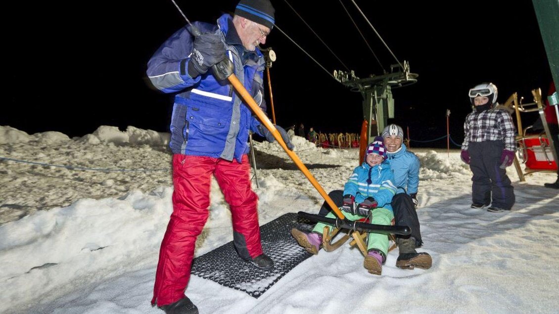 Ein Skilehrer hilft zwei Kinder auf einem Schlepplift in einem verschneiten Gebiet. Es ist Nacht und die Umgebung ist mit Schnee bedeckt. | © Mirja Geh