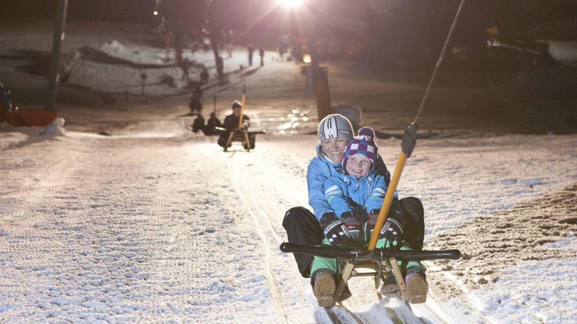 Zwei Kinder fahren gemeinsam auf einem Schlitten den verschneiten Hang hinunter. Die Nachtbeleuchtung sorgt für eine festliche Atmosphäre. | © Mirja Geh