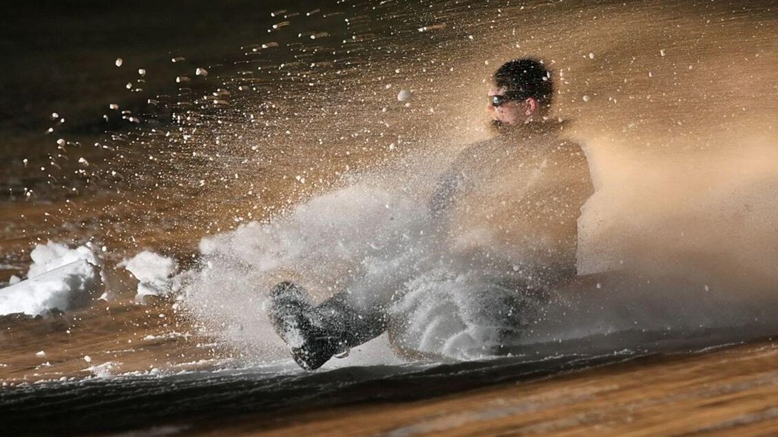 Ein Mann fährt mit einem Schlitten einen Hang hinunter. Schnee spritzt um ihn herum in der Luft. | © Mirja Geh