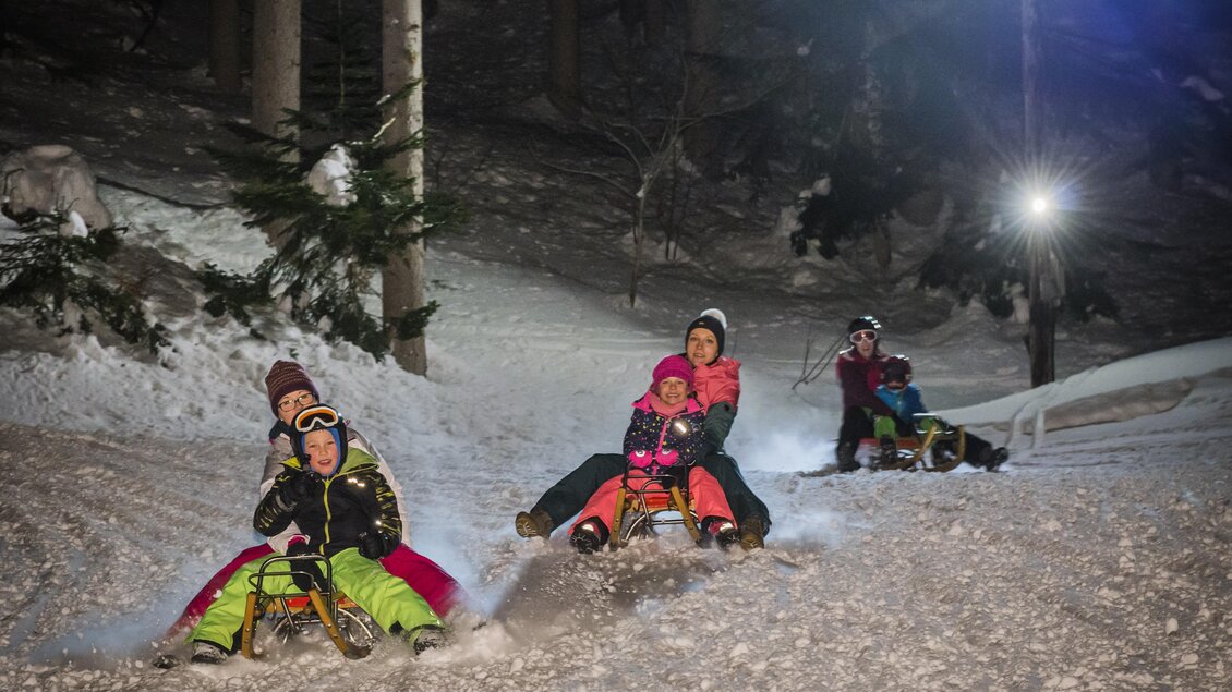 Eine Gruppe von Menschen, die nachts im Schnee rodeln. Die Atmosphäre ist fröhlich und winterlich mit leuchtenden Lichtern im Hintergrund.
