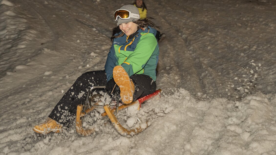 Eine Person fährt mit einem Schlitten den Schnee hinab und hat Spaß. Im Hintergrund ist eine weitere Person zu sehen.
