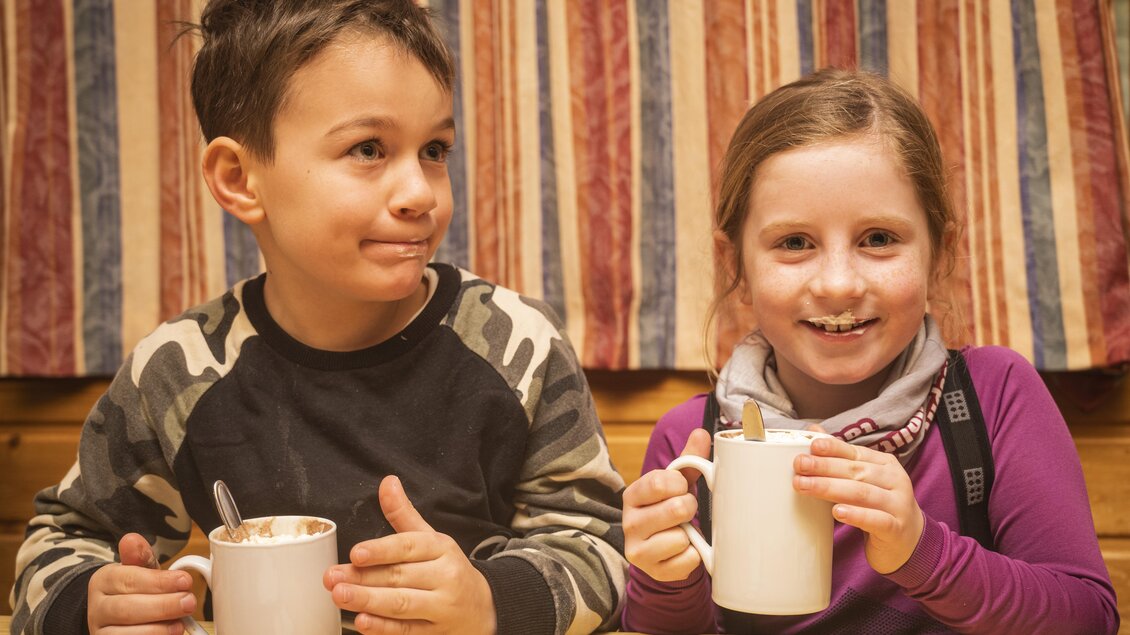 Zwei Kinder sitzen an einem Tisch und halten Tassen mit Getränken. Sie lächeln freundlich und scheinen die gemeinsame Zeit zu genießen.