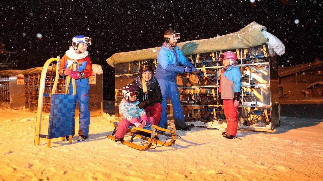 Eine Gruppe von Menschen steht im Schnee und bereitet sich auf das Schlittenfahren vor. Es schneit leicht, und im Hintergrund ist ein Schlittenanhänger zu sehen. | © Hans-Peter Steiner
