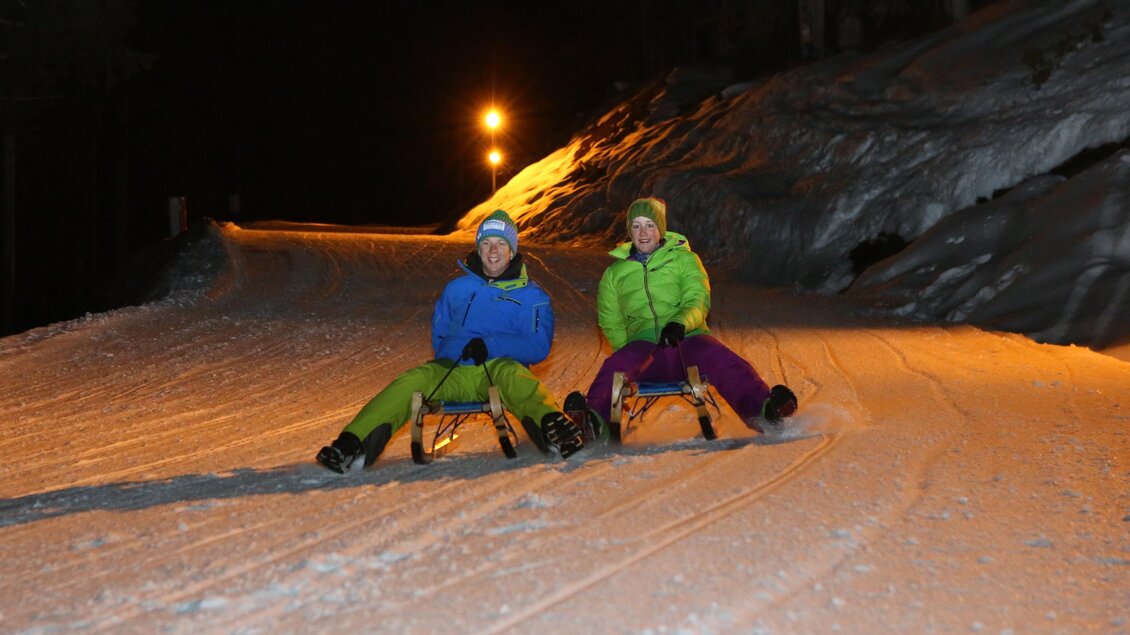 Zwei Personen sitzen in bunten Wintersportanzügen auf Schlitten im Schnee. Die Nacht ist mild erleuchtet von Straßenlaternen. | © Hans-Peter Steiner