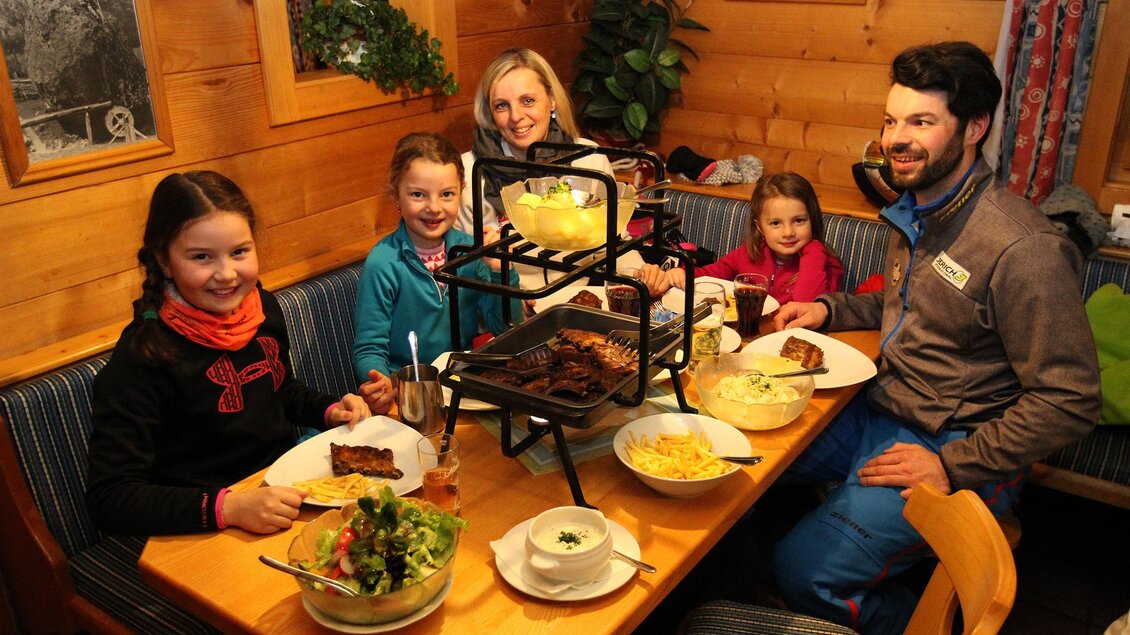 Eine Familie sitzt an einem Tisch in einer gemütlichen Holzhütte und genießt ein gemeinsames Essen. Auf dem Tisch sind verschiedene Gerichte und eine Grillstation zu sehen. | © Hans-Peter Steiner